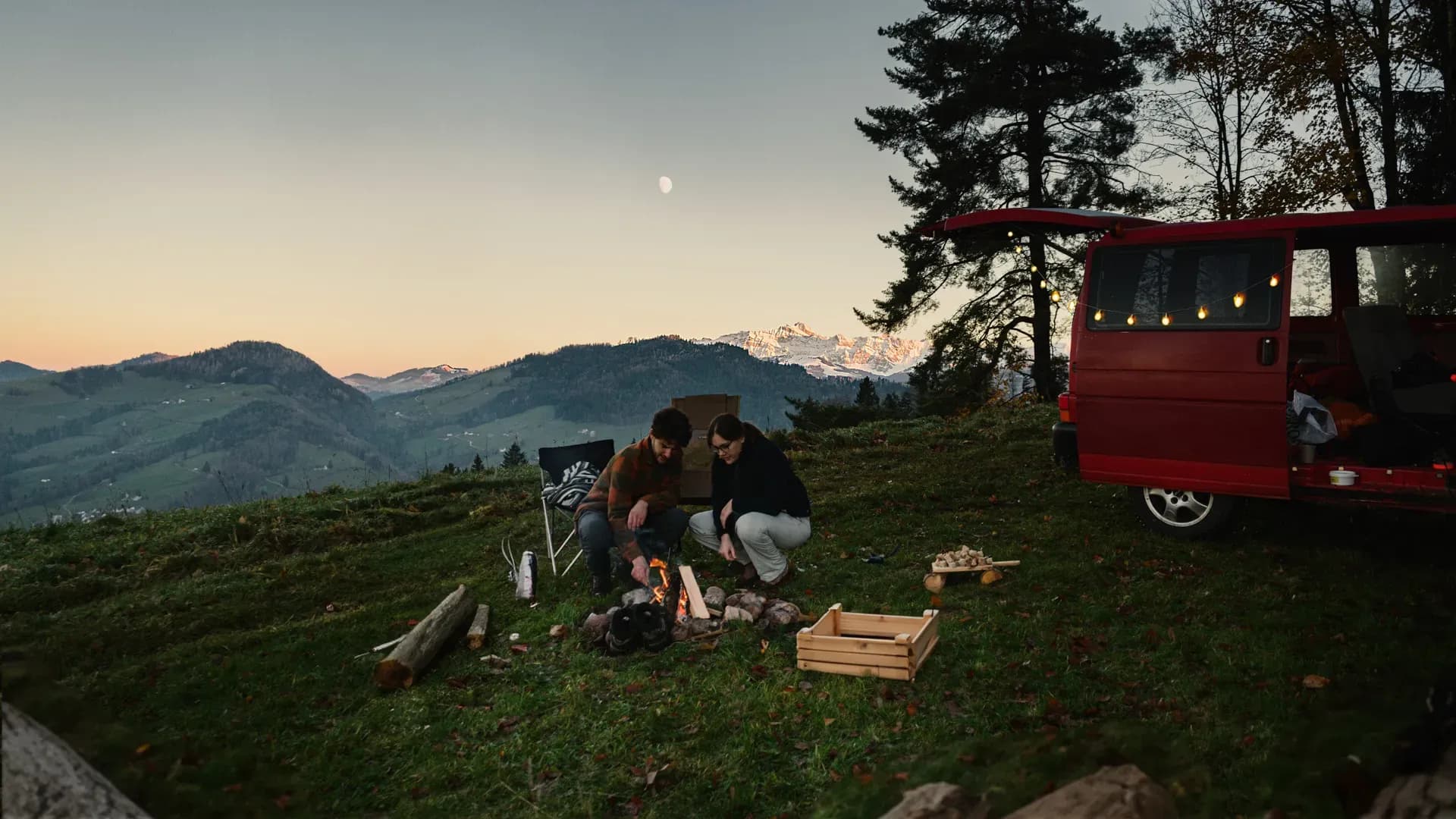 zwei Camper machen ein Lagerfeuer auf einem Nomady Camp bei Mondschein auf einer Waldlichtung vor ihrem Campervan